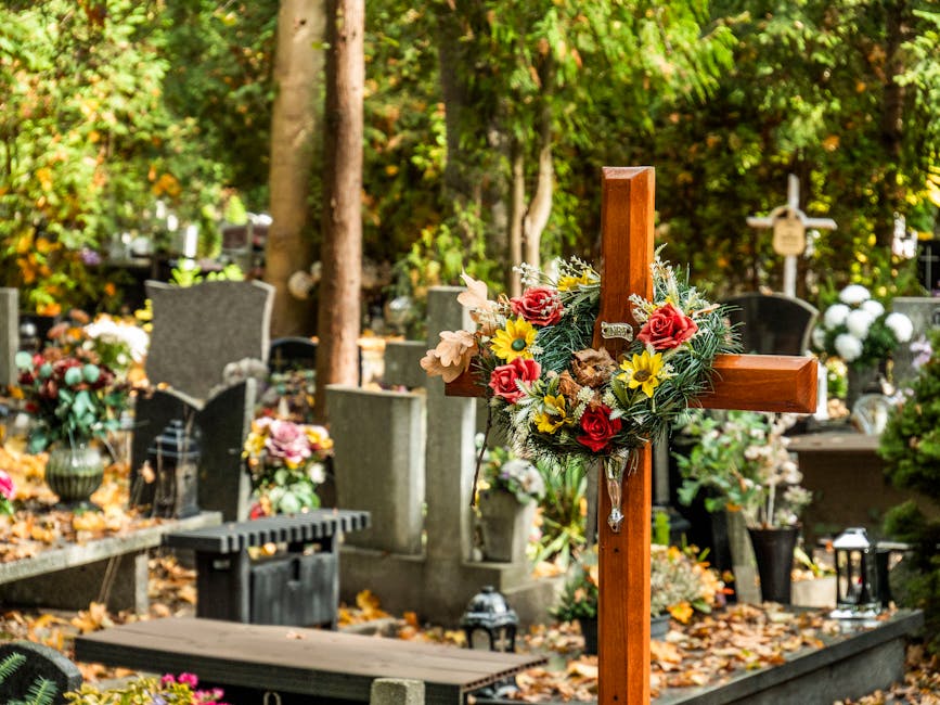 A wooden cross decorated with a floral arrangement featuring red roses, yellow sunflowers, white lilies, and green foliage, set in a cemetery surrounded by various gravestones and memorials. The flowe