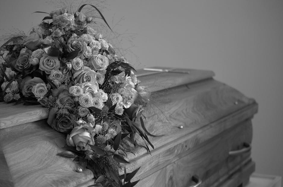A large, elegant bouquet of fresh flowers resting atop a wooden casket, featuring an array of roses in various sizes and shades, complemented by greenery and delicate fillers. The bouquet is wrapped w