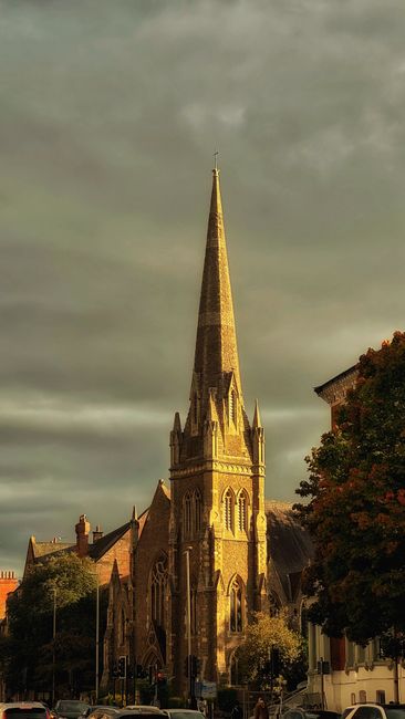 This image features a tall, Gothic-style church with a pointed spire set against a cloudy sky. The church is constructed from light-colored stone, with intricate architectural details including arched