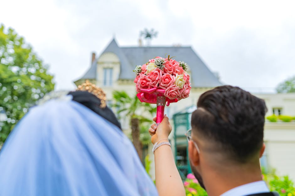 A floral bouquet arranged with pink and cream roses, featuring small white filler flowers and lush green foliage, is held upright by a person wearing a silver bracelet. The bouquet is wrapped with pin