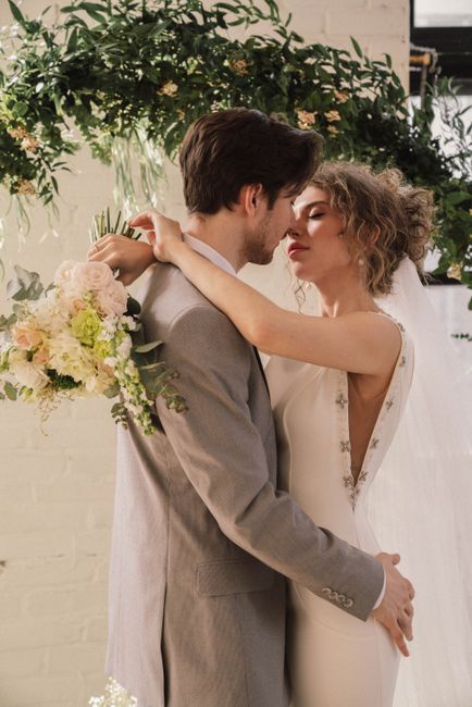 A bride and groom share a tender moment indoors, with the bride embracing the groom and gazing into his eyes. The bride has curly, styled hair and wears a white wedding dress with delicate embroidery 