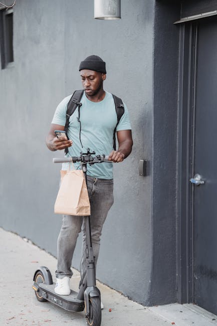 A young man with dark skin, wearing a black beanie, light blue t-shirt, and gray jeans, stands outdoors beside a gray wall near a black door, holding a smartphone in his right hand while resting his l